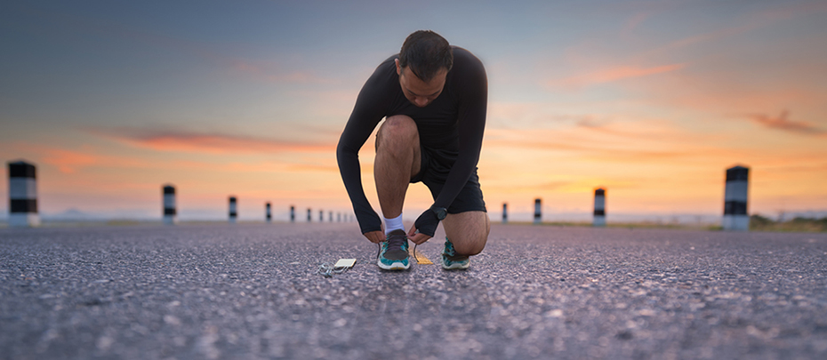 A man in activewear kneels down to tie his shoe on a paved road.