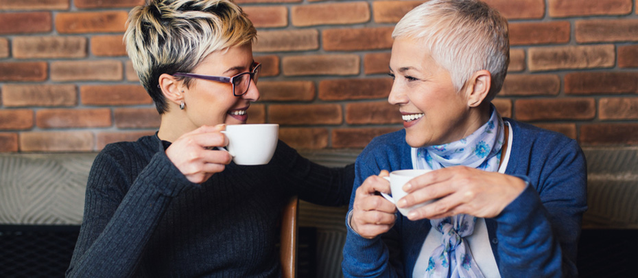 Two women smile at each other while holding mugs at a table in a restaurant.