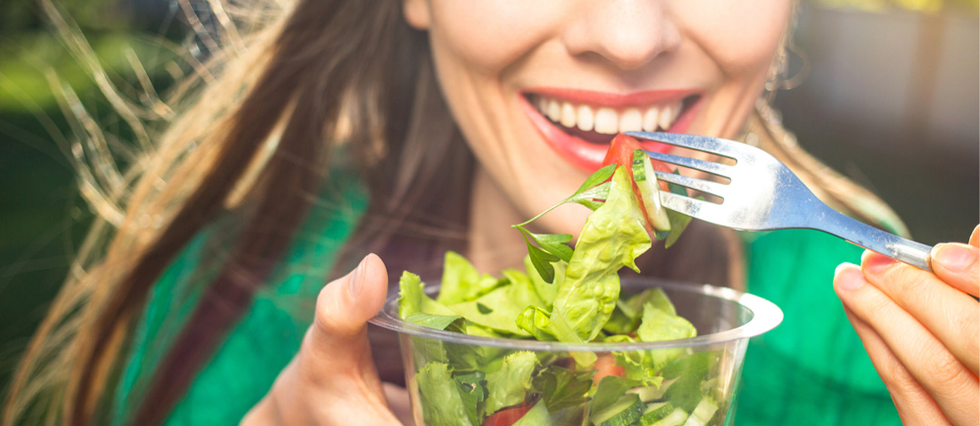A smiling woman lifts a forkful of salad to her mouth.