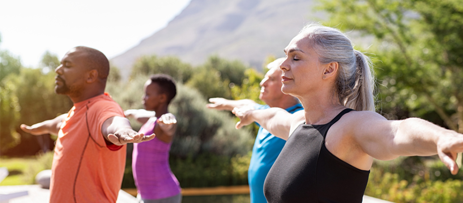 A group of people stands with their eyes closed and arms stretched out in a T pose outdoors on a sunny day.