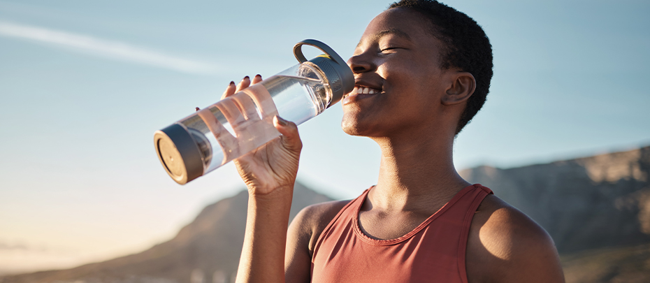 A person smiling and drinking water from a clear bottle with a mountain in the background