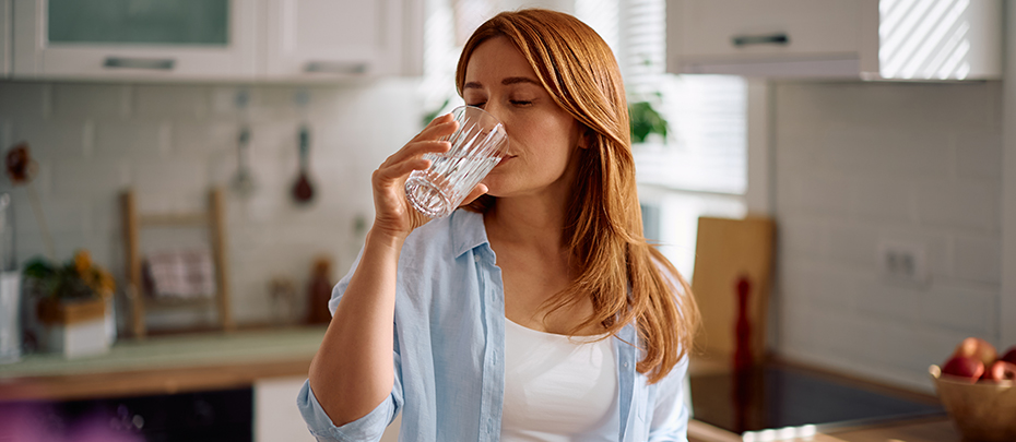 A woman drinks a glass of water standing in her kitchen.