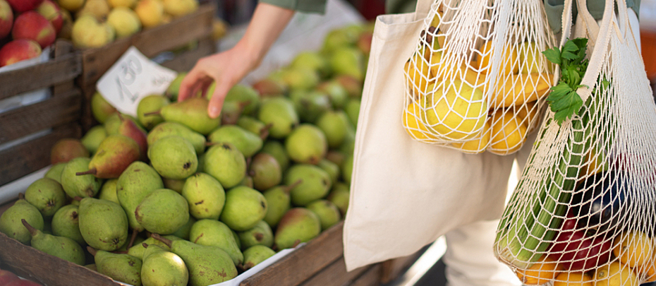 A person holding grocery bags of fruits and vegetables while standing in front of a fruit stand