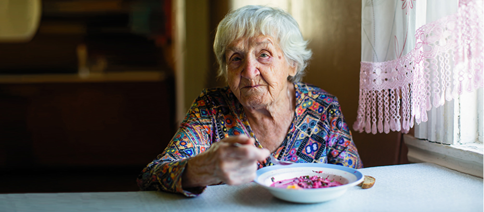 An older woman sits at a table in front of a bowl of food.
