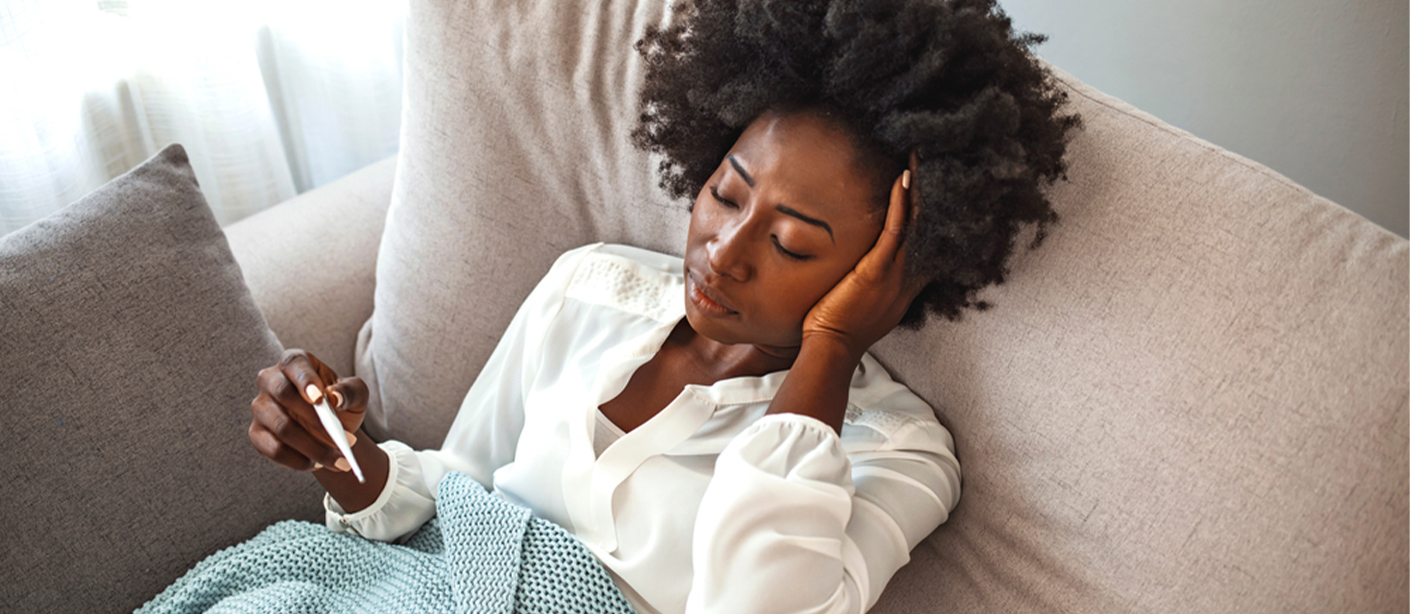 A woman lies under a blanket on the couch with a tissue on her lap, reading a thermometer.