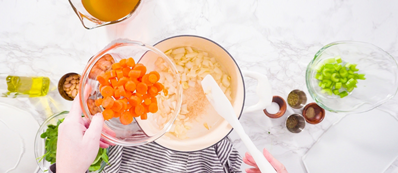 Person adds chopped carrots to a pot of white bean and vegetable soup.