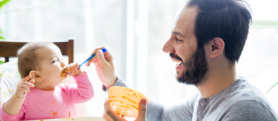 A man feeds solid foods to a baby sitting in a highchair.