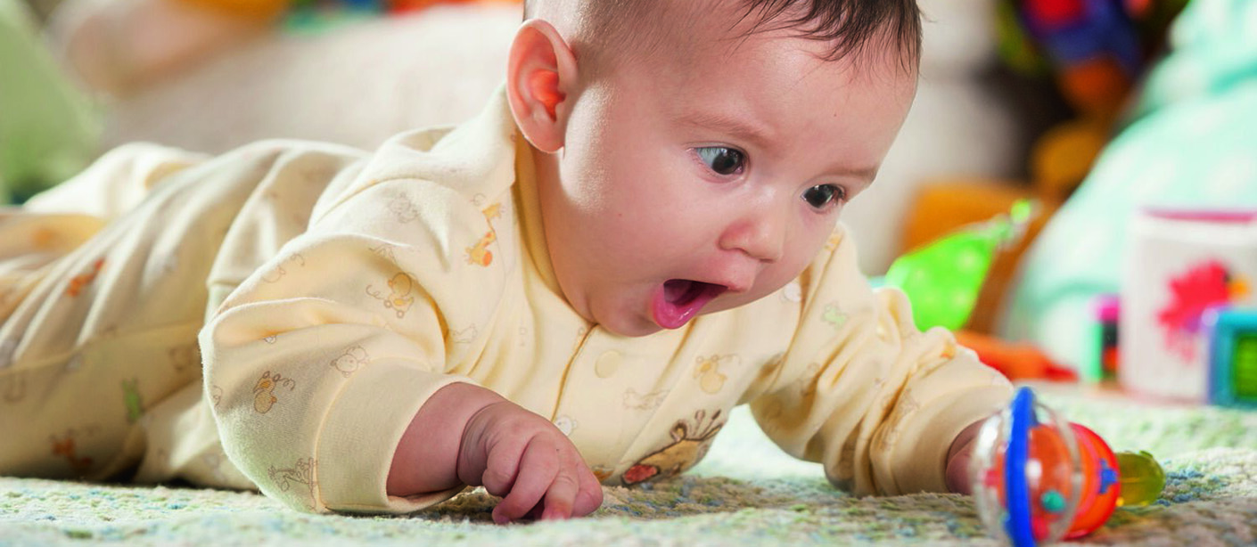 Baby on tummy looking at a rattle on the floor with wide eyes and an open mouth
