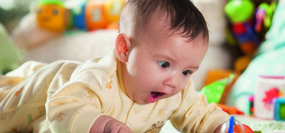 Baby on tummy looking at a rattle on the floor with wide eyes and an open mouth