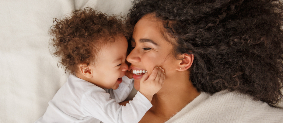 A smiling mother and baby lie face to face on a bed.