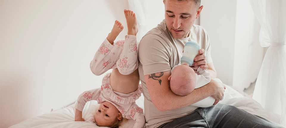 Man gives an infant a bottle while navigating feeding changes and toddler in pajamas plays on the bed behind him.