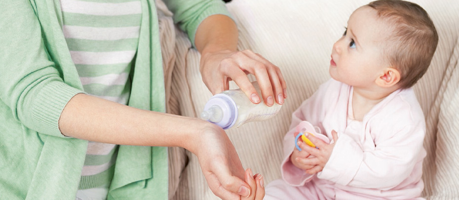 A person tilts a baby bottle toward the inside of their wrist while a baby looks on.
