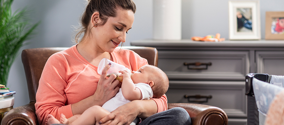 A woman bottle-feeds an infant in a chair.