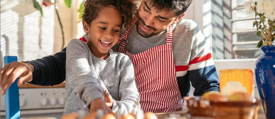 A smiling child reaches for a carton of eggs while their father stands behind them wearing an apron.